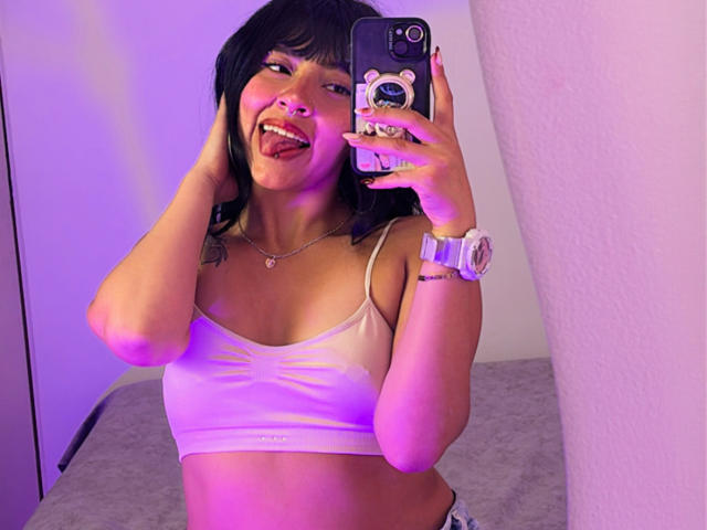 MiaPresscot tirando uma selfie com iluminação roxa, com a língua de fora, vestindo um top esportivo branco e um relógio, segurando um celular com um adesivo de urso..