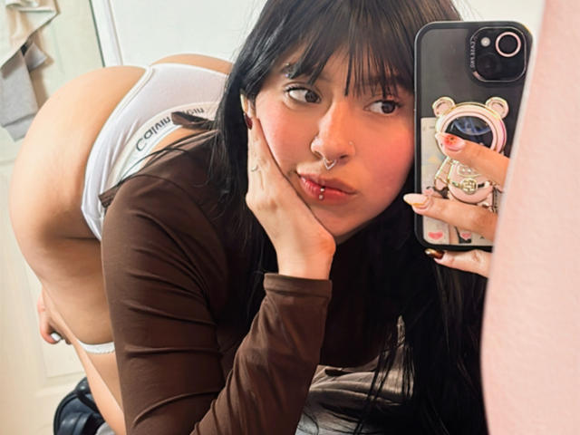 MiaPresscot mit schwarzen Haaren, braunem Langarmshirt, weißen Unterhosen, gebeugt, wobei ihr Hintern zu sehen ist, macht ein Selfie mit ihrem Handy und einer Handyhülle, mit Nasenring und rosafarbenem Lippenstift..