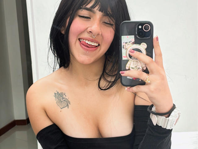 MiaPresscot, con cabello negro, lengua afuera, piercing en la nariz y una blusa negra con un hombro descubierto, sosteniendo un teléfono con una funda con forma de oso, mostrando un tatuaje de corazón en el pecho derecho, sonriendo..