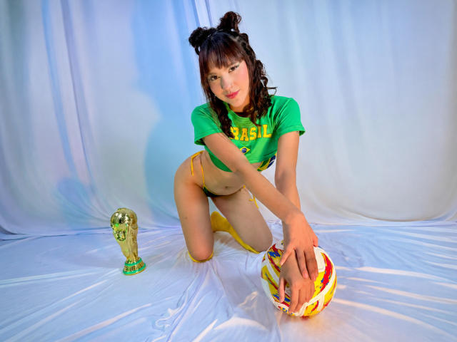 HanaJoud met krullend zwart haar, een groene top, een gele string en sokken, knielend op een wit laken, met een kleurrijke bal in haar handen, naast de gouden FIFA-trofee, kijkend naar de camera..