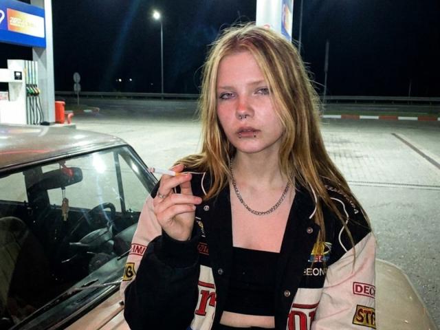 SocorroZamzow with long blonde hair, nose ring, and chain necklace, wearing a black crop top and sports jacket, smoking near a car at a night-time gas station.