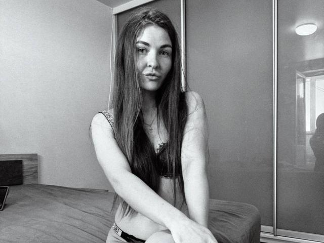 LumiEmma has long, straight hair, small tits in a black bra, and a slight smile. She's sitting on a bed, arms crossed, in a black-and-white photo.
