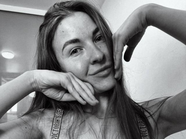 Black-and-white selfie of LumiEmma with long hair, resting chin on hands, wearing a tank top with strap details, slight smile, freckles visible, looking relaxed.