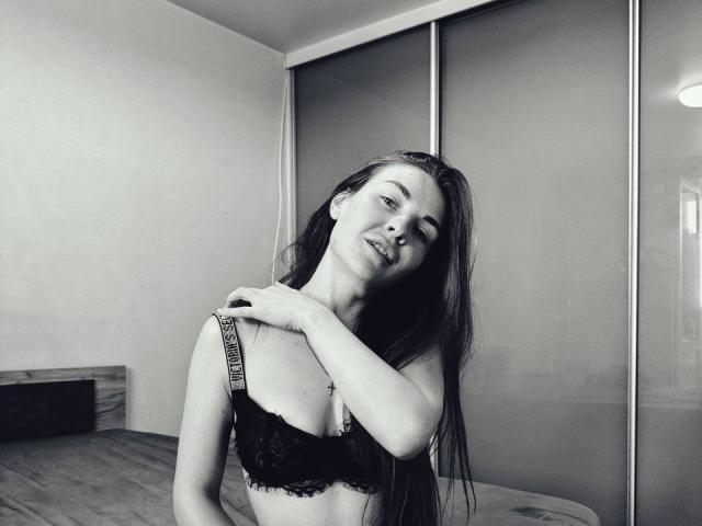 LumiEmma has long dark hair, wearing a black lace bra, her hand on her shoulder, slightly tilted head, and a seductive smile in a black-and-white photo.