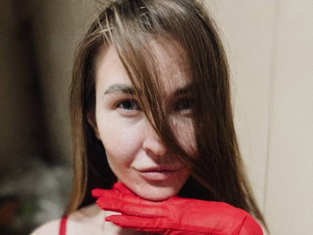 LumiEmma with long brown hair, red gloves, and a subtle smile, covering her neck, looking at the camera with a slightly mischievous expression. Her tits are not visible.