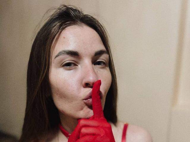 LumiEmma with long brown hair, light skin, and subtle makeup, sucking on a red fingerless glove, looking directly at the camera. Boobless, no visible genitals, just the glove.