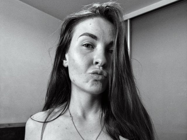 Black and white photo of LumiEmma with long, messy hair, pouty lips, and a necklace, showing her bare shoulders. She has a slight smile and a confident gaze.