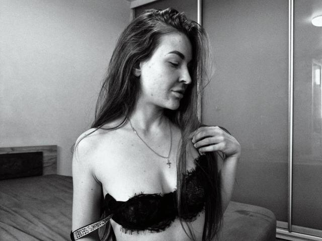Black-and-white photo of LumiEmma with long hair, wearing a lace bra, touching her hair, a cross necklace, and a tattoo on her upper arm, eyes closed, smiling softly.