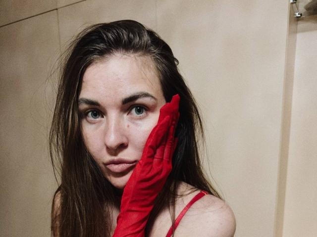 LumiEmma with wet brown hair, blue eyes, and red glove covering her cheek, wearing a red spaghetti strap, looking serious. Her tits are slightly visible.