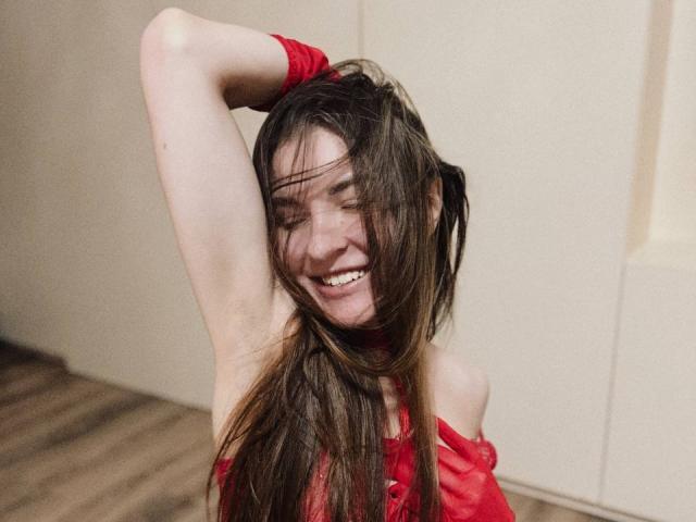 LumiEmma with long brown hair, red off-shoulder top, arm raised, smiling, eyes closed, hair in face, showing armpit, wooden floor, white wall.