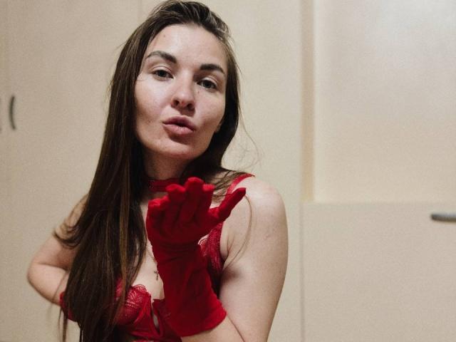 LumiEmma with long brown hair, wearing red lace lingerie and matching red gloves, pouting slightly, her tits visible, hands near her face, in a soft-lit indoor setting.