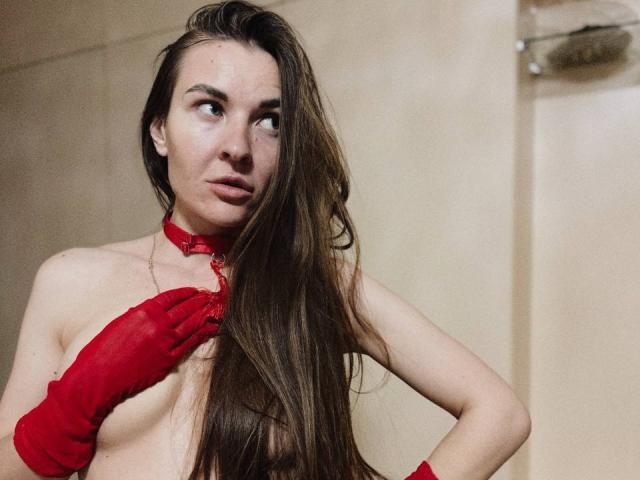 LumiEmma with long brown hair, red choker, and red gloves, covering her small tits with one hand, looking to the side, in a dimly lit room.