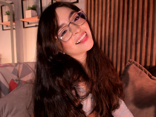 Eva McAllister mit langem, gewellten, dunklen Haar, Brille und einem süßen Lächeln, die ihre Zunge herausstreckt. Sie trägt ein hellgraues Oberteil und sieht glücklich und verspielt aus..