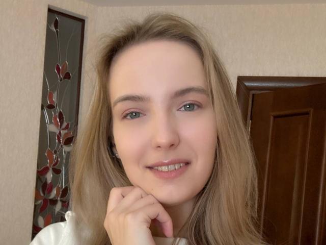 Nicole Spritzer, avec ses longs cheveux blonds, ses yeux verts et un léger sourire, repose son menton sur sa main, montrant ses dents. Elle porte un haut blanc et a l'air adorable..