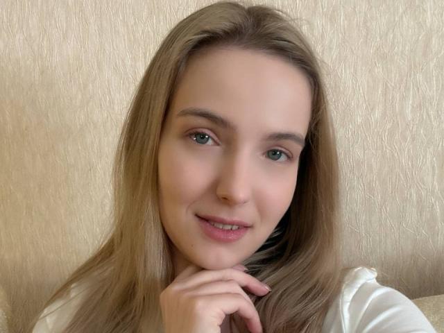 Nicole Spritzer ha lunghi capelli biondi, occhi verdi e un leggero sorriso, con una mano che tocca il mento. Indossa una maglietta bianca, ed è carina e attraente..