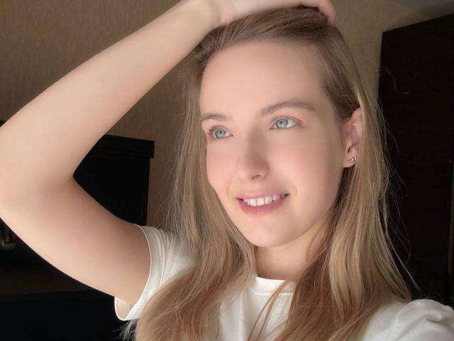 Nicole Spritzer, com cabelo loiro, olhos azuis e camisa branca, sorrindo, braço direito levantado, mão no cabelo, mostrando um piercing na orelha, e pele macia..