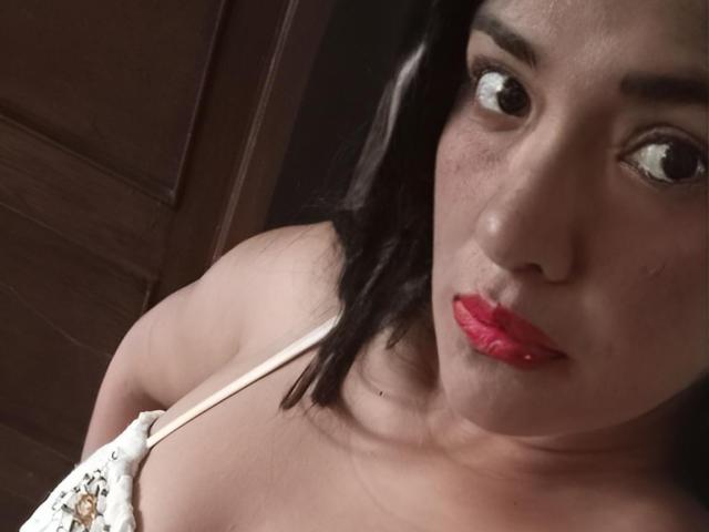 ArianithaXox, con piel media, cabello negro, lápiz labial rojo y una blusa blanca con tirantes finos, que muestra su parte superior del pecho, mirando a la cámara con una expresión ligeramente fruncida..