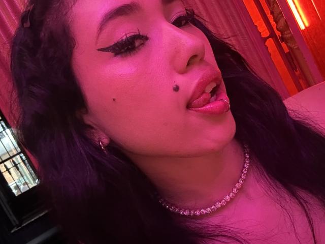 NashiroSakuraii has dark wavy hair, black winged eyeliner, a pierced tongue, a lip piercing, and multiple facial moles. She wears a diamond choker and pinkish-purple lighting.