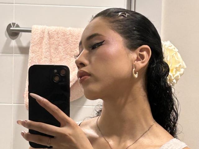 NashiroSakuraii with wet black hair, gold earrings, and bold eyeliner, taking a selfie in a bathroom, pouting slightly, holding a black phone, pink towel in the background.