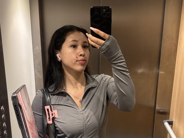 Nashiro Sakurai, met een gemiddelde borstmaat, draagt een grijze ritsjas en maakt een selfie in een lift. Ze houdt een zwarte telefoon, een roze tasriem en witte oordopjes vast..
