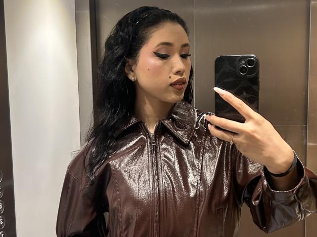 NashiroSakuraii with long black hair, wearing a shiny brown leather jacket, taking a selfie in a mirror with a black phone, having full, dark lips and a small beauty mark.