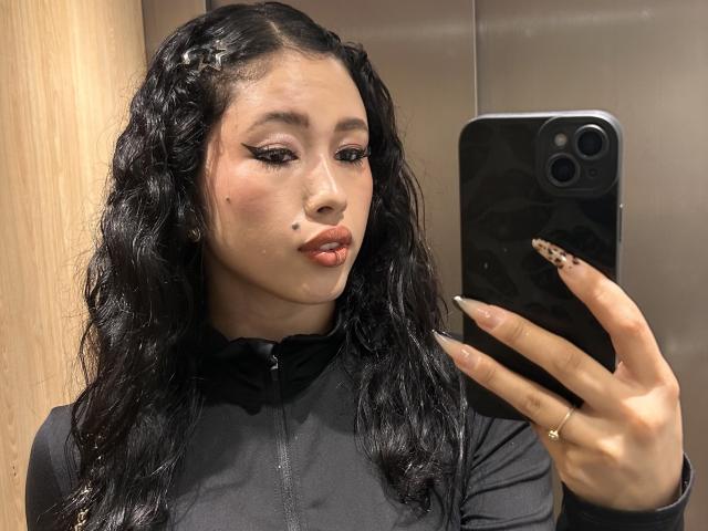 Nashiro Sakurai, met krullend zwart haar, gekleed in een zwarte ritsjas, met een telefoon in haar hand, die haar luipaardprint nagels laat zien, en een klein sproetje op haar rechterwang, in een spiegelzelfportret..
