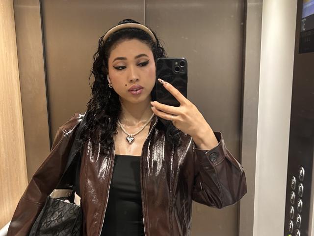 NashiroSakuraii has curly black hair, brown leather jacket, black tube top, silver necklace, and holds a black phone in an elevator mirror selfie, with a serious expression.
