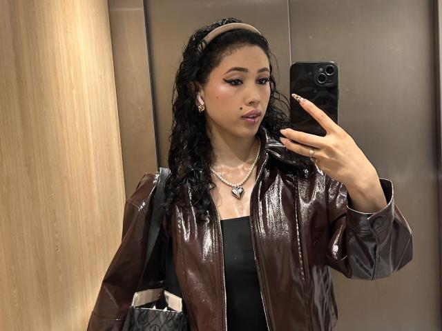Nashiro Sakurai, met golvend zwart haar, draagt een glanzend bruin leren jack, een zwarte top en een parelketting, en maakt een spiegelzelfportret in een lift, terwijl ze een zwarte telefoon vasthoudt..