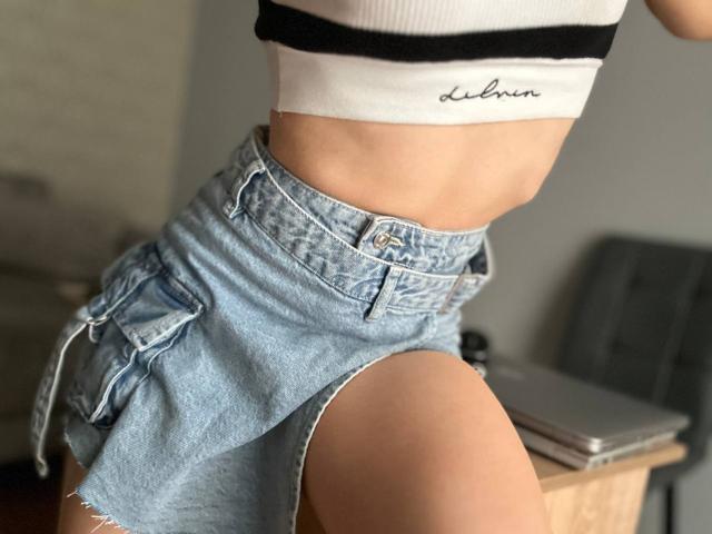 FortyLeride in a white crop top, showing a slim waist, and wearing a high-waisted, light-blue denim skirt with a front pocket, sitting with one thigh raised.
