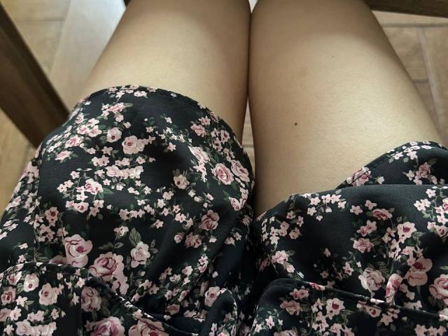 De benen van FortyLeride, gekleed in een bloemenjurk, zwarte stof met roze rozen, licht gespreid, waarbij bleke huid en een kleine, donkere moel op de rechter dij zichtbaar zijn..
