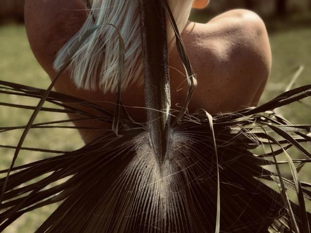 FortyLeride's back with long, silver hair, holding palm fronds, centered on a thick, vertical plant stem, sunlight highlighting the texture.