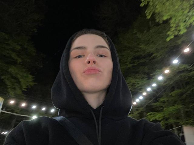 ChristiniaMarian with pouty lips, wearing a black hood, staring into the camera, soft lighting, green tree branches in the background, string lights overhead, dark night, casual selfie.