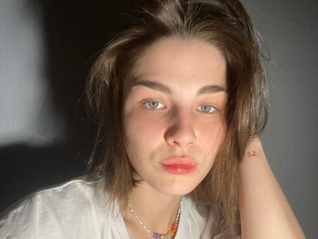 ChristiniaMarian has pale skin, green eyes, and slightly parted pinkish lips. She's wearing a white shirt with a colorful necklace. She's got a red tattoo on her right wrist.