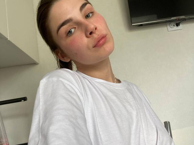 ChristiniaMarian with fair skin, green eyes, and brown hair in a ponytail, wearing a white shirt, pouting slightly, looking at the camera. Her tits are not visible.