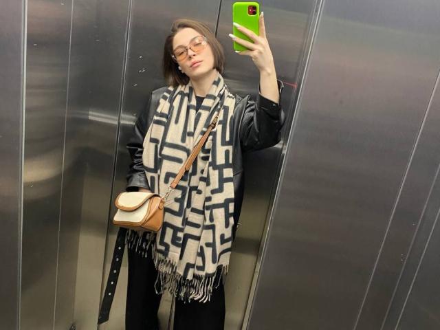 ChristiniaMarian taking a mirror selfie in an elevator, wearing a black leather jacket, geometric scarf, black pants, glasses, and a tan crossbody bag, holding a green phone.