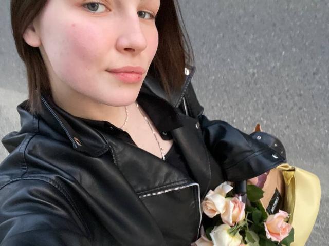 ChristiniaMarian with light skin, brown hair, black leather jacket, black shirt, holding yellow bag with pink and white roses, slight smile, gray pavement background.