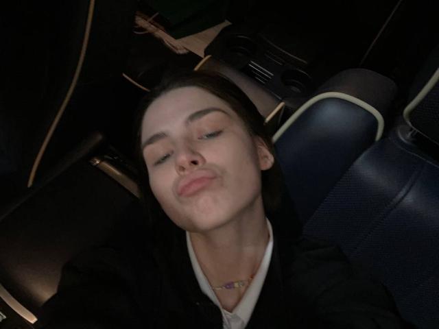 ChristiniaMarian has her eyes closed, a small smirk on her face, and her head tilted back in a car seat. She's wearing a black jacket and a white shirt.