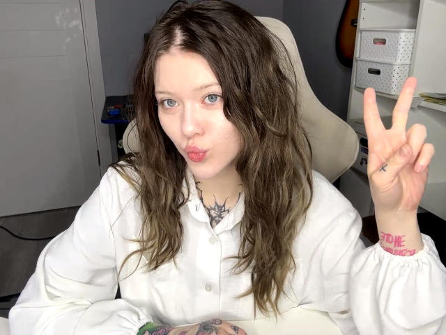 MeliaRinaldo with wavy brown hair, pale skin, and blue eyes, wearing a white button-up, pouting lips, flashing a peace sign, tattoo on neck, and "LOVE" tattoo on wrist.