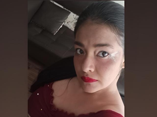 ArianithaXox met donker haar, rode lippenstift en witte oogmake-up, kijkt omhoog met grote ogen, en draagt een rode top met kleine gouden stipjes, in een zwak verlichte kamer..