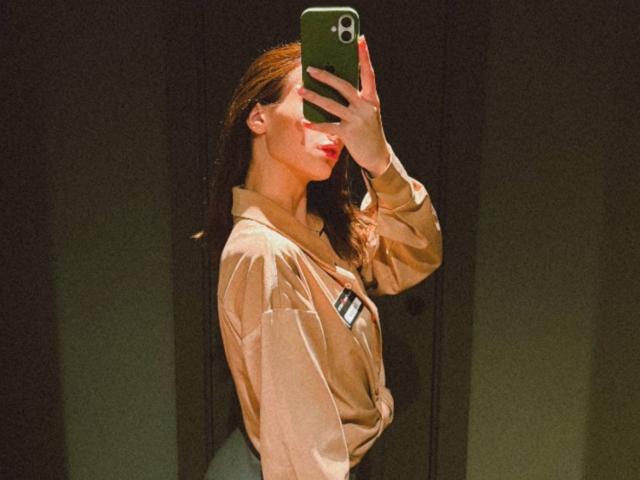 MistieVakas with red lipstick, holding green phone, wearing beige shirt tied at waist, showing side profile, medium tits, brown hair, mirror selfie, dim lighting.