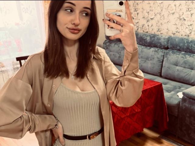 MistieVakas, con i capelli castani, indossa una camicia beige sopra un vestito grigio a coste e una cintura nera, sta scattando un selfie allo specchio, mostrando il seno e una collana con una croce..