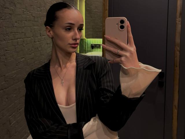 MistieVakas, con el cabello peinado hacia atrás, vistiendo un blazer negro a rayas, un vestido blanco y un collar con una cruz, se está tomando un selfie frente al espejo con un iPhone, mostrando sus senos..