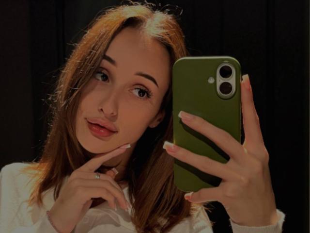 MistieVakas, con cabello castaño claro, sosteniendo un teléfono verde, tiene un dedo en el mentón, lleva una camisa blanca, tiene labios rosados y una sonrisa encantadora..