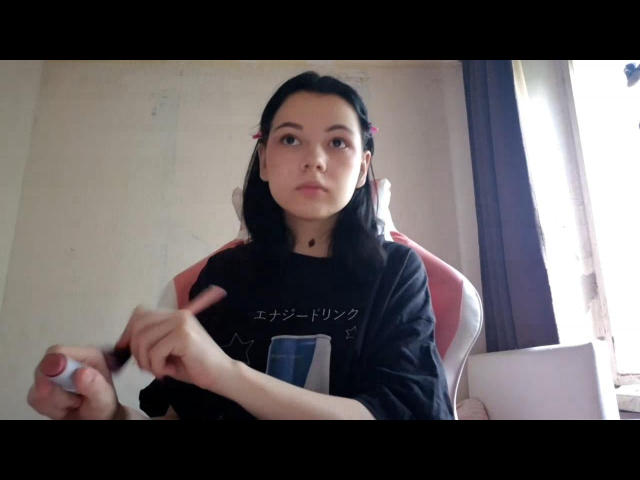 AaronKolesnik has black hair, pink hair clips, and a black shirt. She's pointing with her right index finger, holding a pen in her left hand.