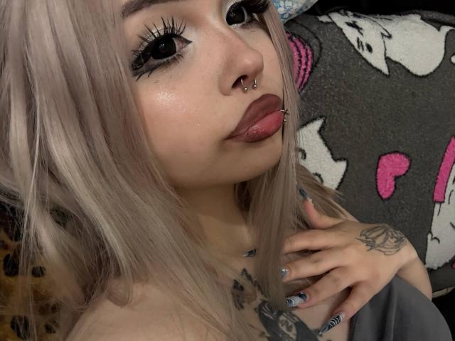 SerinaSindlinger with platinum blonde hair, black eyeliner, nose ring, and lip piercing, tongue slightly out, hand on chest, tattoos visible, dark pink and white blanket in background.