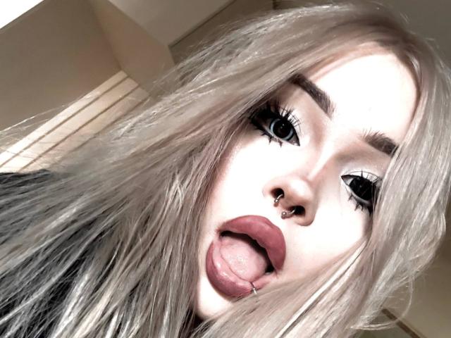 SerinaSindlinger has pale skin, long blonde hair, and a nose piercing. She's sticking out her tongue with pink lipstick and her eyes are wide with heavy eyeliner.