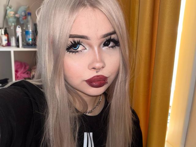 SerinaSindlinger has long blonde hair, blue eyes, thick black eyelashes, red lipstick, and a nose ring. She's wearing a black shirt and a silver chain.