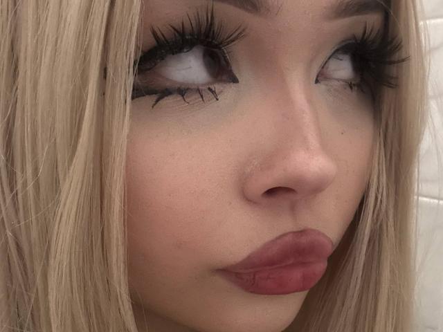 SerinaSindlinger's face with blonde hair, thick black eyelashes, and pink lipstick, looking slightly up with a neutral expression. Her nose is small and her skin is fair.