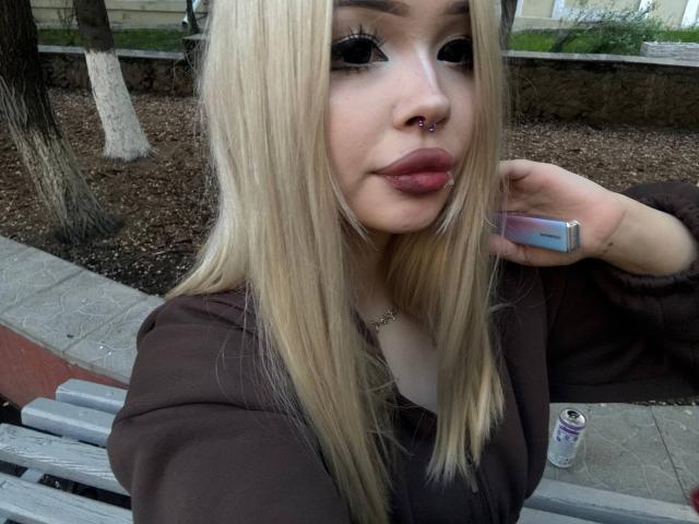 SerinaSindlinger with long blonde hair, nose ring, and pink lipstick, wearing a brown top, holding a phone, can, and wooden bench in the background. Her tits are visible.