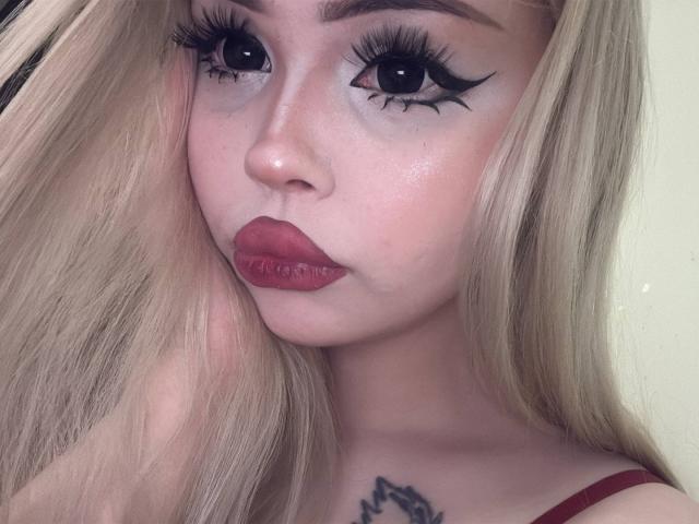 SerinaSindlinger has blonde hair, dark eyeliner, and red lipstick. Her eyes are big and black, and she has a tattoo on her chest. Her tits are small.
